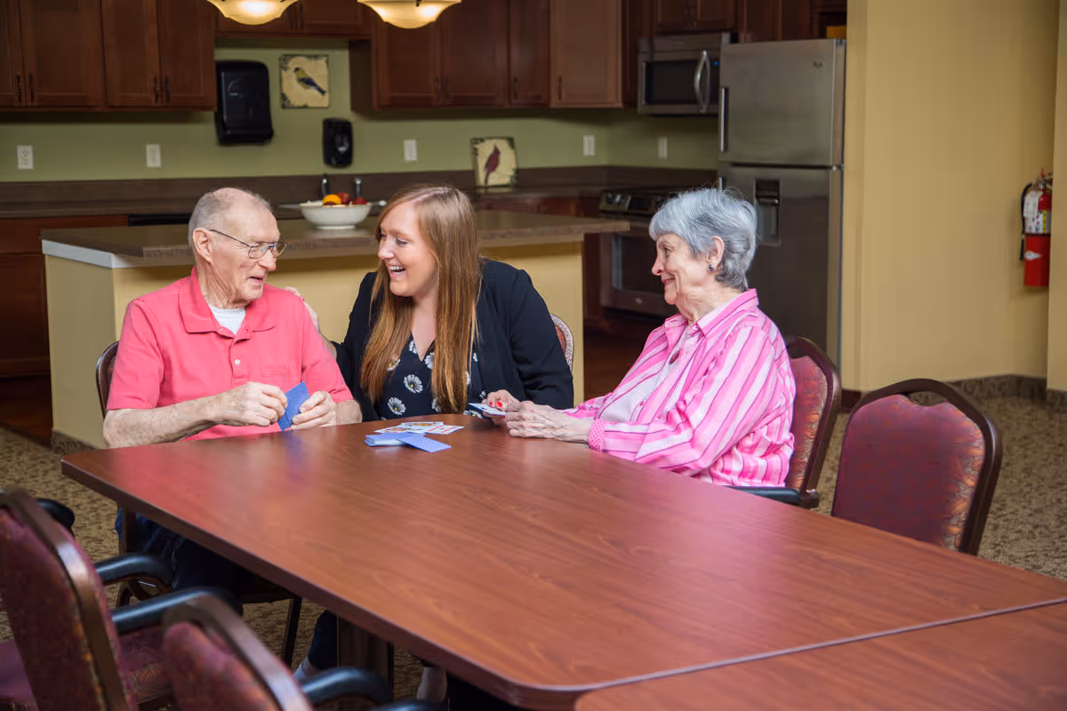 Two elderly individuals and a younger woman sitting around a wooden table playing cards in a room with a kitchen area in the background. The elderly man is wearing a red shirt, the younger woman has long hair and is smiling, and the elderly woman is wearing a pink striped shirt.
