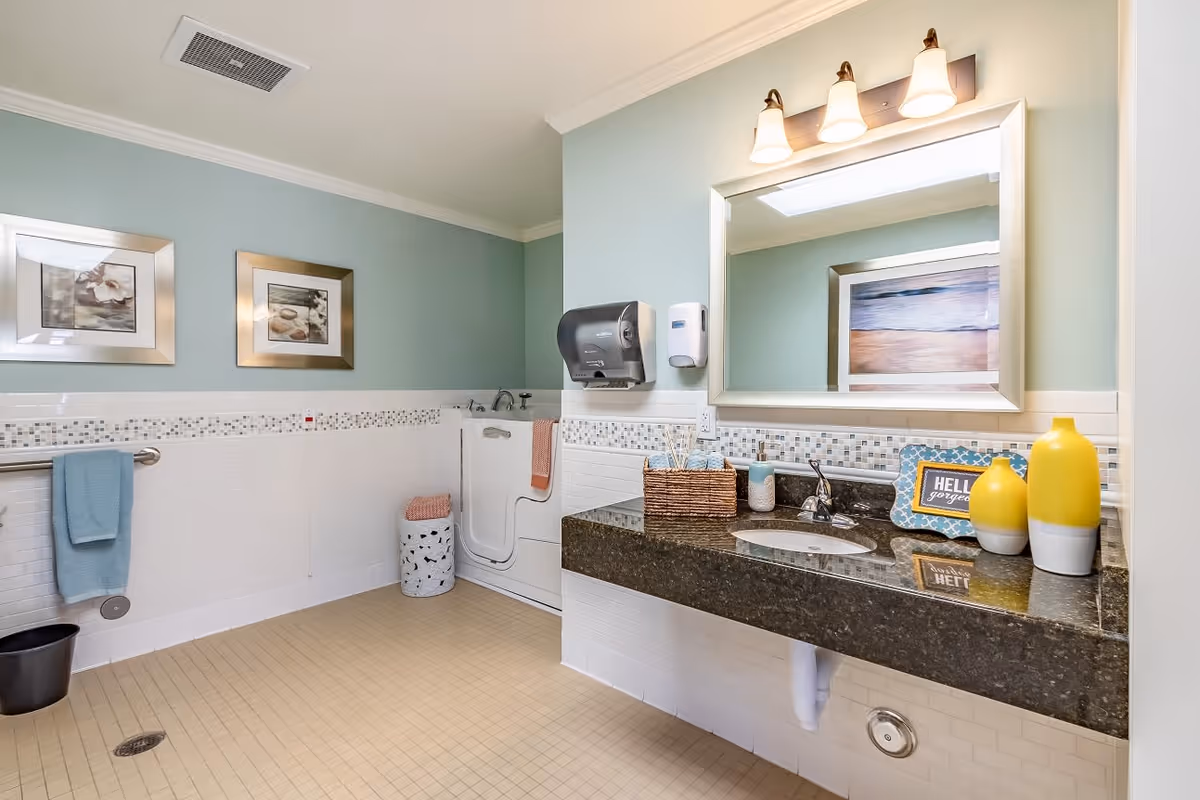 A clean and spacious bathroom with light blue walls, a large mirror above a dark granite countertop with a sink, two yellow and white decorative vases, a basket with toiletries, and a soap dispenser. There is a walk-in bathtub with a door, a towel rack with a blue towel, two framed pictures on the wall, and a beige tiled floor.