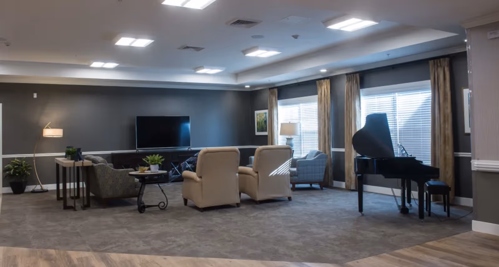 A spacious and well-lit living room area with a large flat-screen TV mounted on a dark gray wall. The room features two beige armchairs, a patterned armchair, a small round table with a plant, a floor lamp, and a grand piano near windows with beige curtains. The floor is carpeted with a wood floor border.