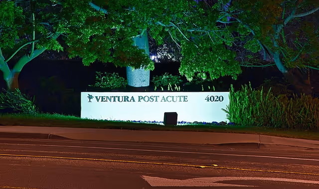 Illuminated roadside sign reading 'Ventura Post Acute 4020' at night with trees and landscaping behind it.