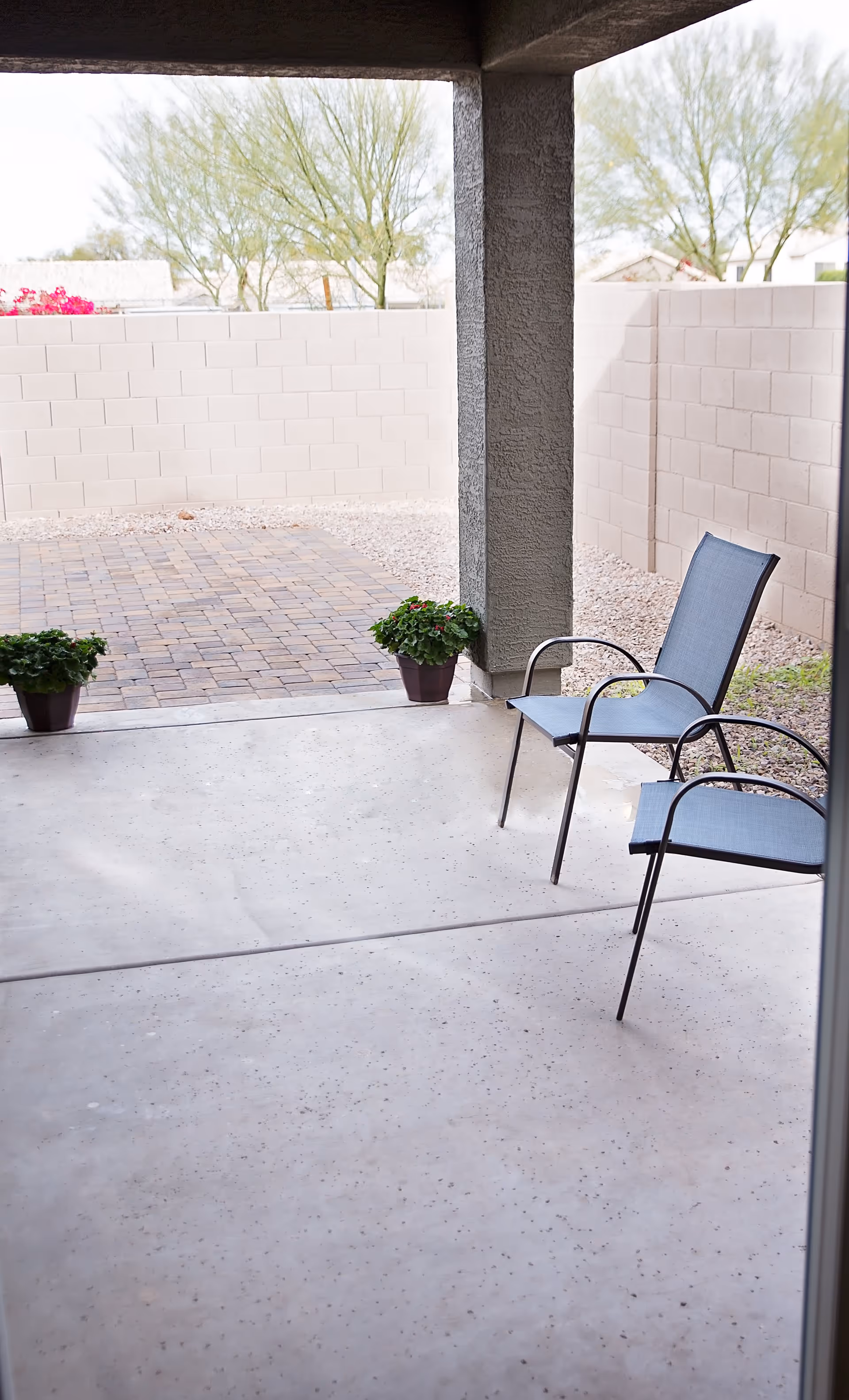 Covered patio area with two blue mesh chairs and two potted plants on a concrete floor, enclosed by a beige brick wall with trees visible in the background.