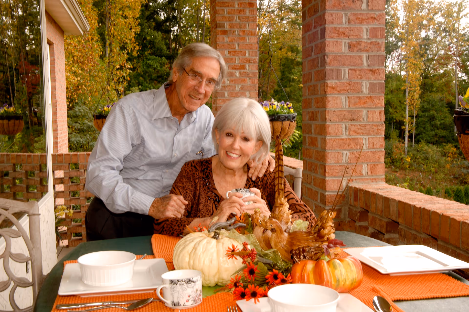 An elderly couple smiling and posing together on a brick patio decorated with autumn-themed centerpiece including pumpkins and flowers. The woman is seated holding a cup, while the man stands behind her with his arm around her shoulder. The table is set with white dishes and orange placemats, and there are hanging flower baskets and trees in the background.
