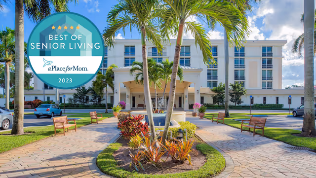 Exterior view of a multi-story senior living facility with a paved walkway, benches, palm trees, and landscaped garden in front. The sky is blue with some clouds. A circular badge in the top left corner reads 'Best of Senior Living aPlaceforMom 2023'.