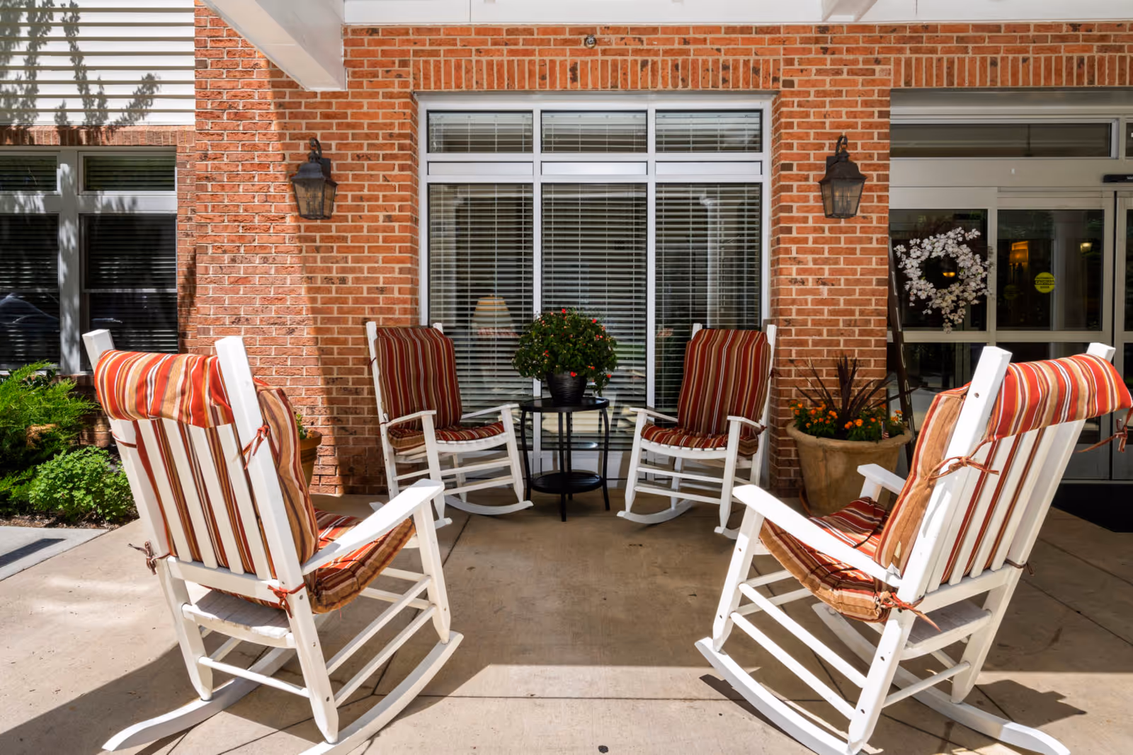 Front entrance seating area with four white rocking chairs with red-striped cushions around a small table in front of a brick wall and windows.