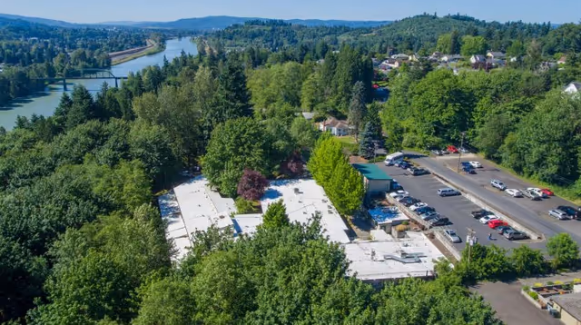 Aerial view of Beacon Hill Rehabilitation facility surrounded by dense green trees, with a river and hills in the background. The image shows the building's white rooftops, a parking lot with several cars, and a nearby road.
