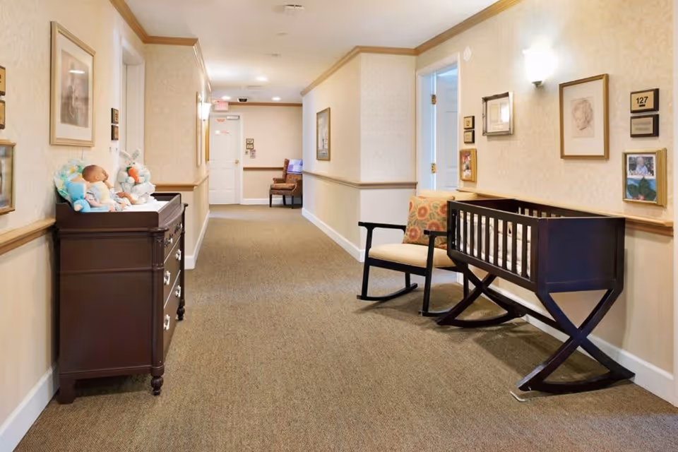 A well-lit hallway in a senior living facility with beige walls and carpeted floor. On the left side, there is a dark wooden dresser with baby dolls and stuffed animals on top. On the right side, there is a dark wooden cradle and a cushioned rocking chair with a decorative pillow. The walls are adorned with framed pictures and room number plaques.