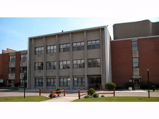 Exterior view of a multi-story senior living facility building with a combination of gray and red brick walls, multiple windows, a small landscaped area with bushes, and a paved walkway leading to the entrance.