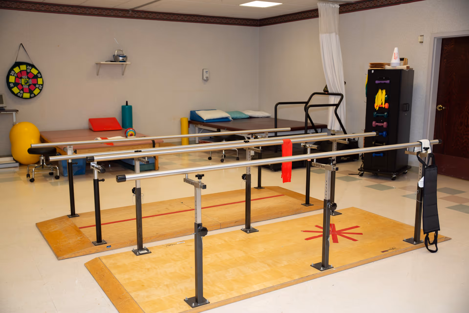 A senior rehabilitation room featuring parallel walking bars on wooden platforms with exercise mats, therapy balls, and equipment along the walls.