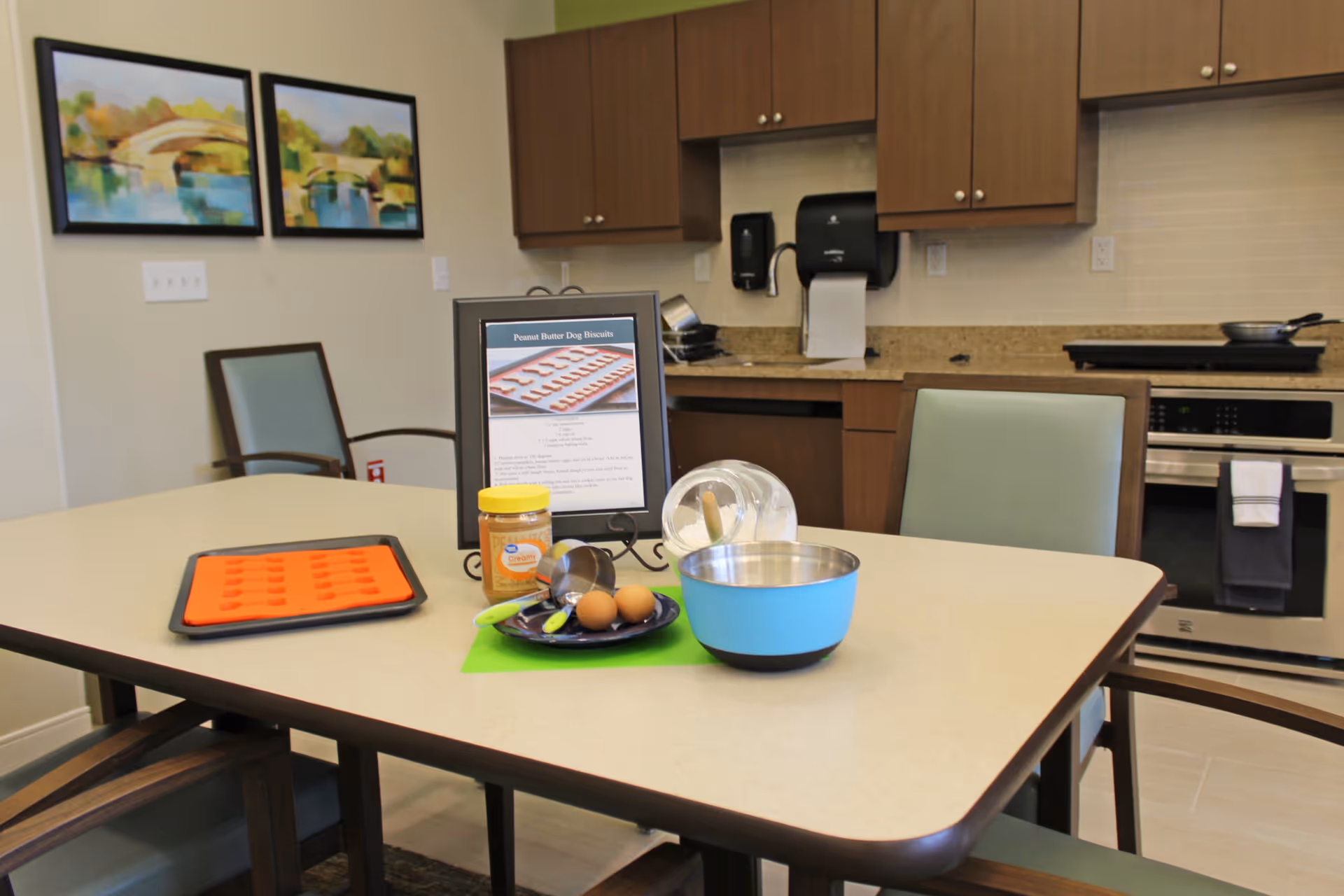 A kitchen area in a memory care facility with a table in the foreground holding baking ingredients including eggs, peanut butter, measuring spoons, a mixing bowl, and a silicone baking mat. Behind the table are wooden cabinets, a stove, and a paper towel dispenser. Two framed paintings hang on the wall.