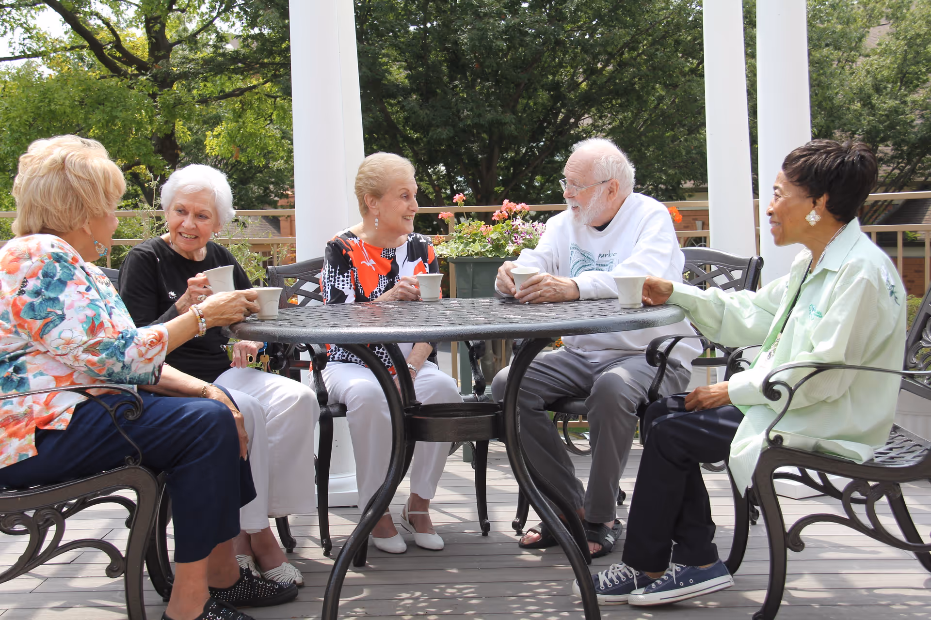 Five elderly people sitting around a round metal table on an outdoor patio, enjoying drinks and engaging in conversation. The setting includes white pillars, green trees, and potted flowers in the background.