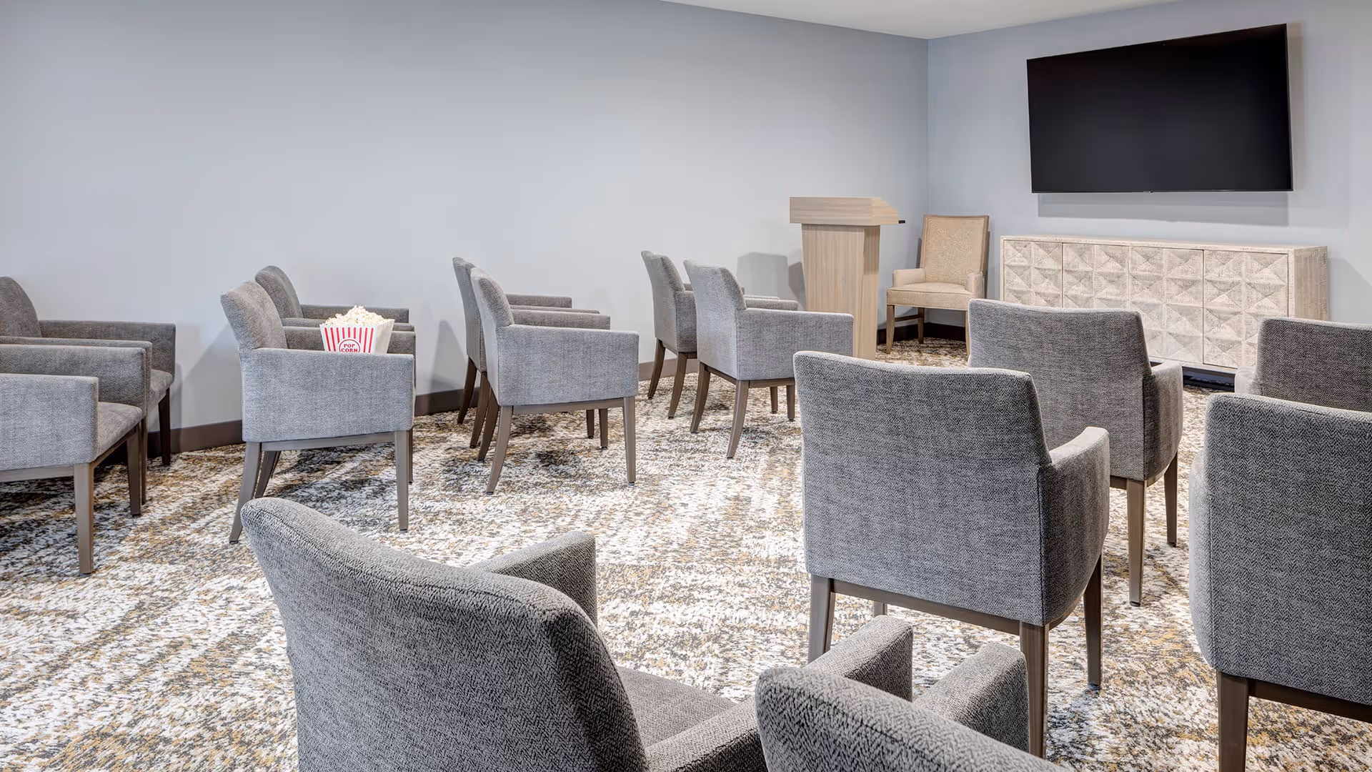A small conference or presentation room with several gray upholstered chairs arranged in rows facing a wooden podium and a large flat-screen TV mounted on the wall. The room has a patterned carpet and light gray walls.
