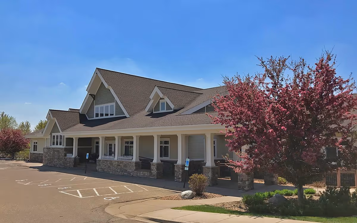 Exterior view of a single-story building with a large covered porch supported by white columns and stone bases. The building has a steeply pitched roof with multiple gables and several windows. In front of the building, there is a parking area with marked handicap spaces. To the right, there is a tree with pink blossoms and some landscaping with bushes and grass. The sky is clear and blue.