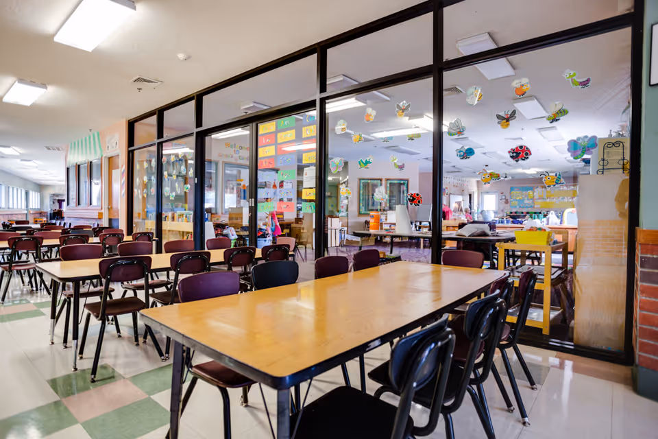 Long indoor dining/activity area with rows of tables and chairs facing a glass-partitioned craft room decorated with paper insects.