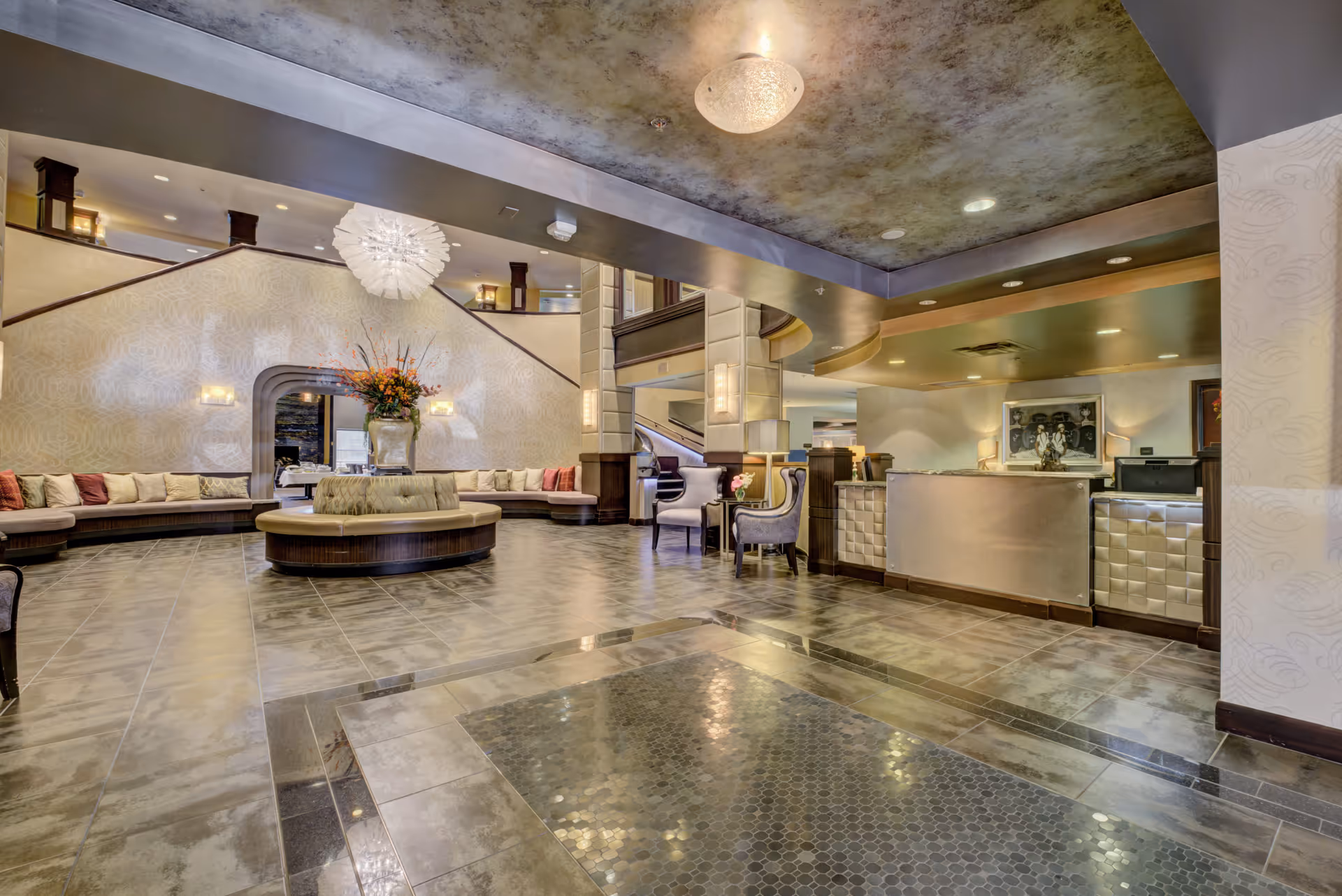 Spacious and elegant lobby area of a senior living facility with a large circular seating arrangement in the center, cushioned benches along the walls, decorative lighting fixtures on the ceiling, a reception desk on the right, and a staircase leading to an upper floor in the background.