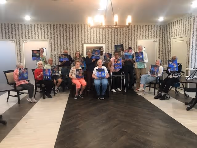 A group of elderly individuals seated and standing in a room holding up artwork featuring a blue and purple design. The room has patterned wallpaper, a chandelier, and several doors in the background. Some individuals are seated in chairs, and one person is using a walker.