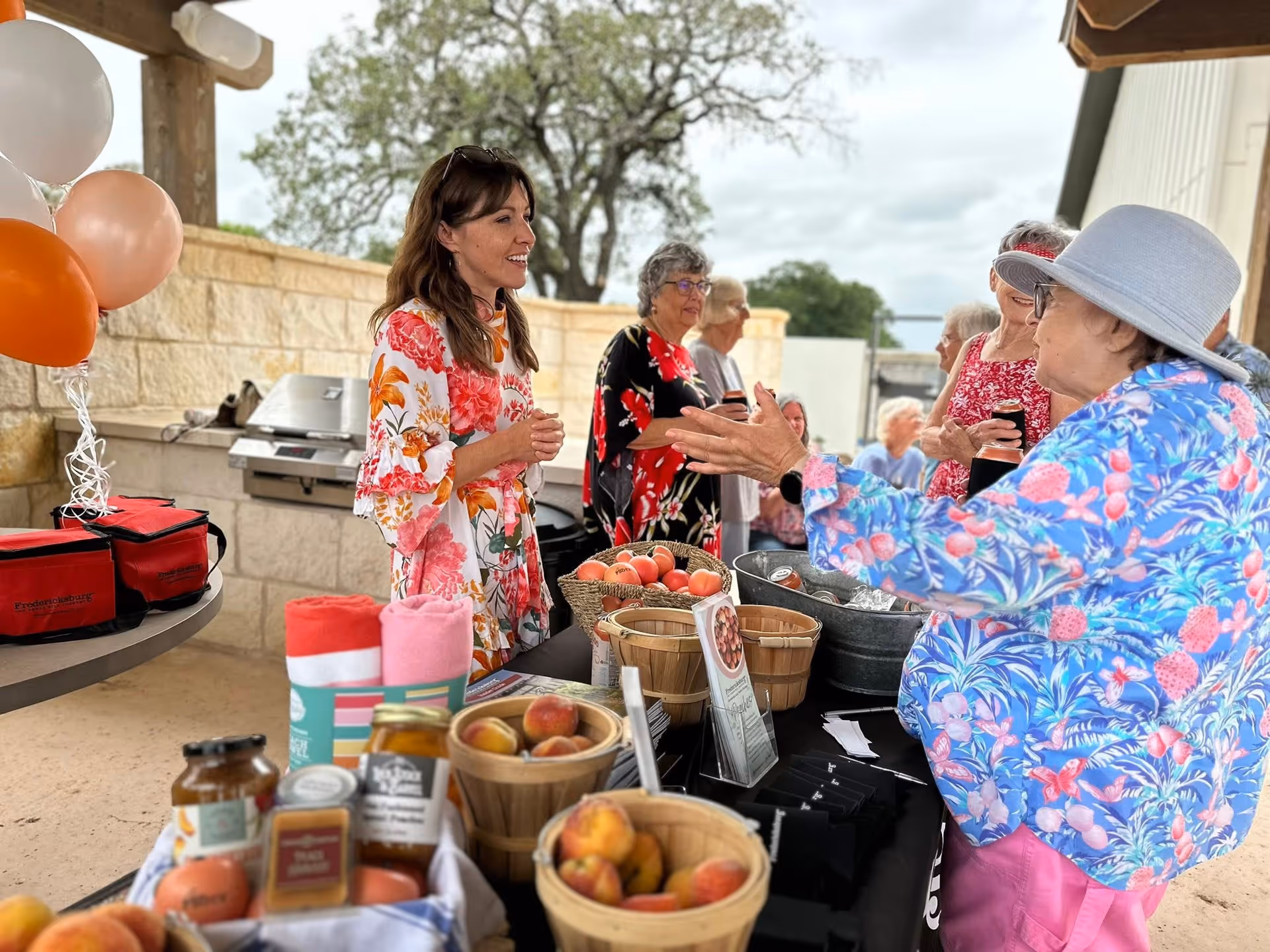 A group of senior women and a younger woman are gathered around a table outdoors at Woodland Cottages Senior Living Fredericksburg. The table is covered with baskets of peaches, jars of preserves, and rolled towels. The women are engaged in conversation, with one woman in a blue floral jacket and hat gesturing animatedly. Balloons are tied to a nearby table, and a grill is visible in the background under a covered patio area.