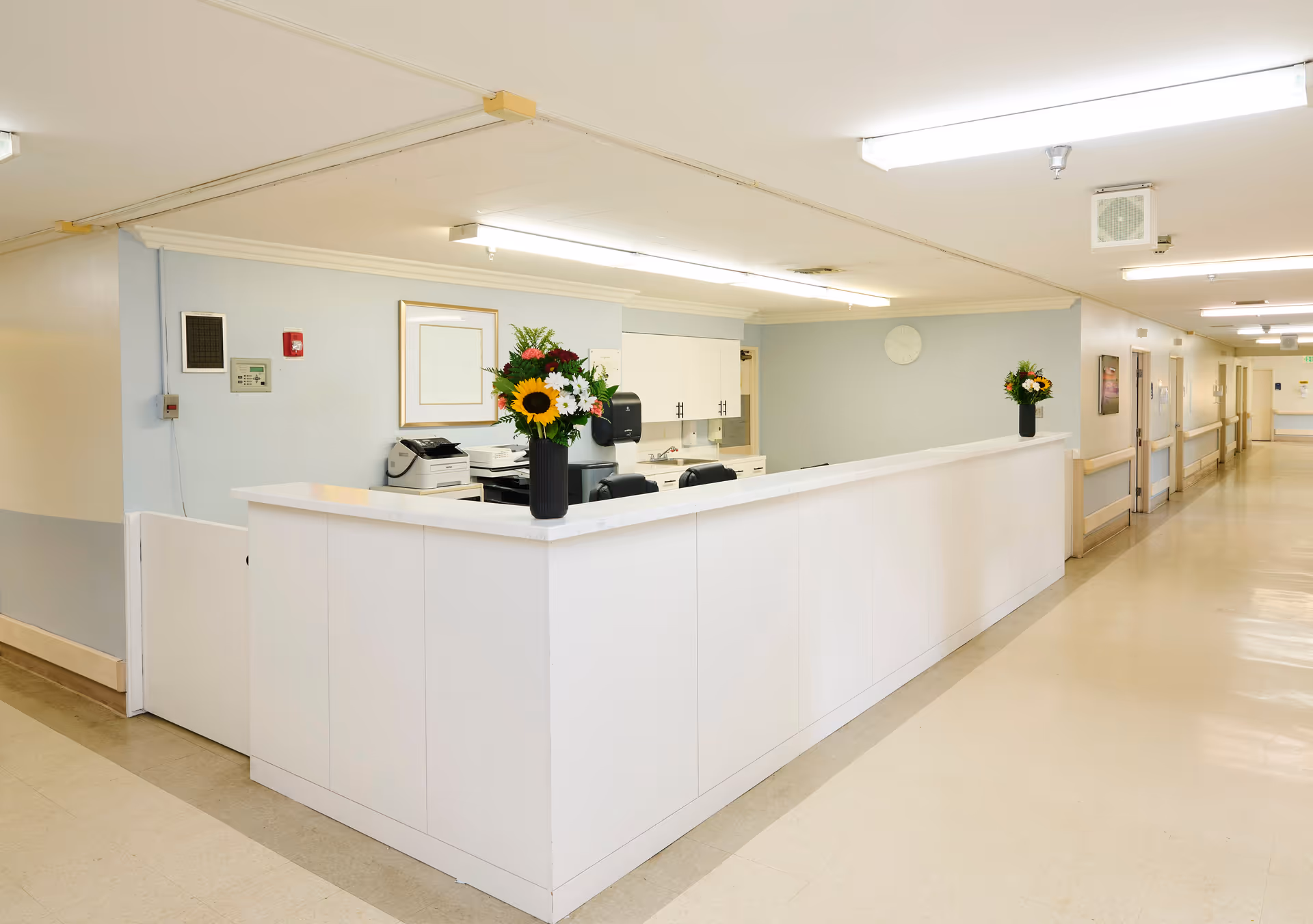 A clean and well-lit nursing station in a senior care facility hallway with a white counter, two vases of colorful flowers, office equipment, and a clock on the wall.