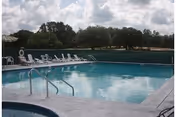 Outdoor swimming pool with metal handrails and a row of lounge chairs beside it, trees and cloudy sky in the background.