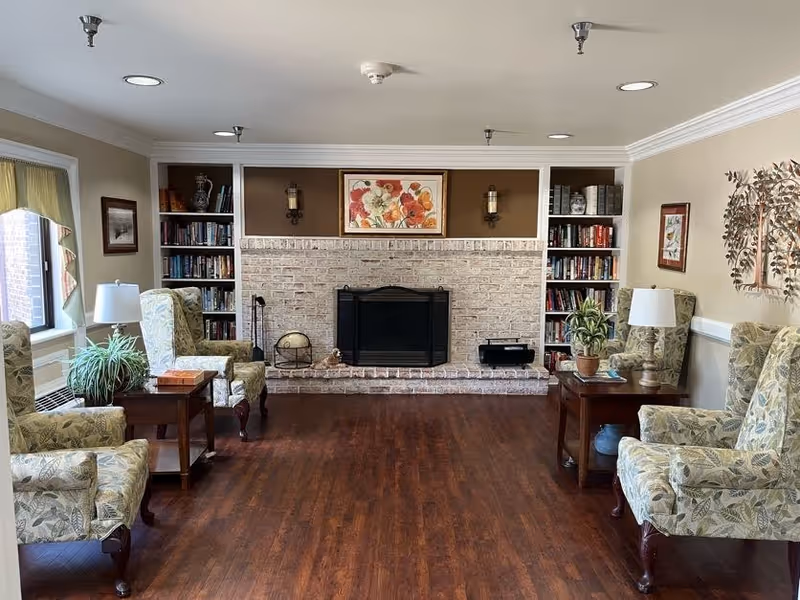 A cozy living room with four floral-patterned armchairs arranged around two wooden side tables with lamps and plants. The room features a brick fireplace with a floral painting above it, built-in bookshelves on either side filled with books and decorative items, and hardwood flooring. There are windows with curtains on the left side and wall decorations on the right.