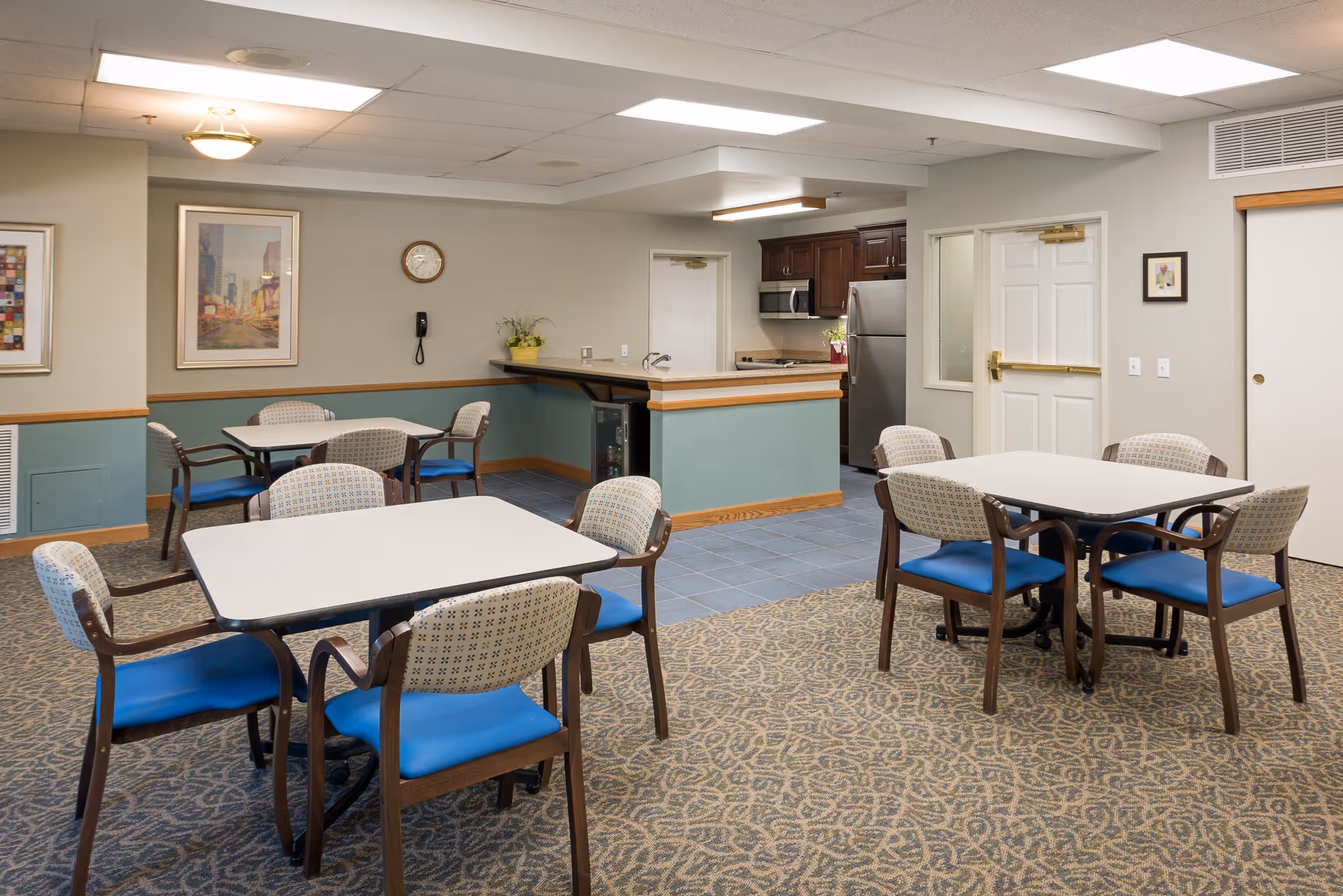 A senior living facility dining area with multiple tables and chairs featuring blue cushions. The room has patterned carpet flooring and a tiled section near a small kitchen area with wooden cabinets, a stainless steel refrigerator, and a microwave. The walls are decorated with framed artwork and a clock, and there is a telephone mounted on the wall.