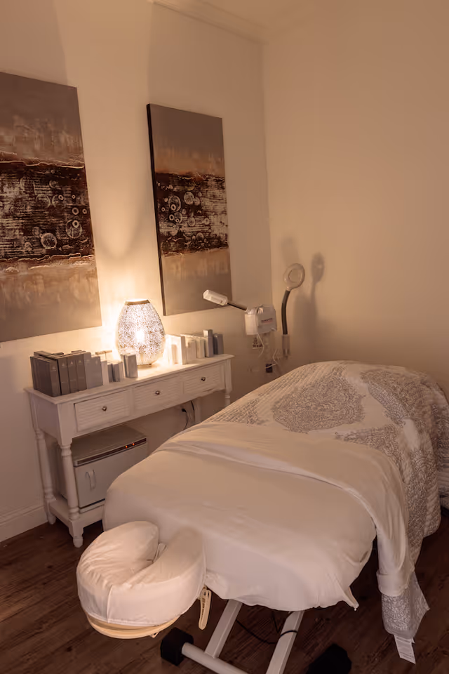 Softly lit treatment room featuring a massage table with face cradle, a bedside console with lamp and products, and abstract wall art.