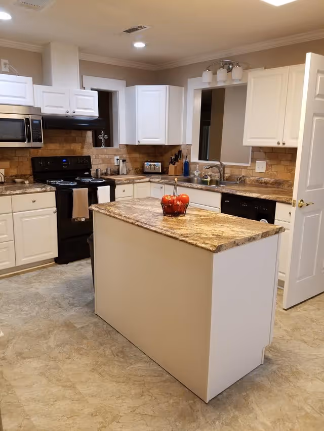 A modern kitchen with white cabinets, a black stove and microwave, a beige and brown granite countertop island with a basket of red apples, and a sink area with a window and light fixture above it. The floor has a light-colored tile, and the walls have a beige tile backsplash.