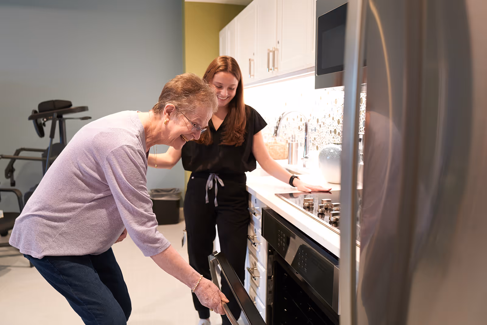 An elderly woman is opening an oven door in a modern kitchen while a younger woman stands behind her smiling. The kitchen features white cabinets, a stovetop, and a stainless steel refrigerator. In the background, exercise equipment is visible.
