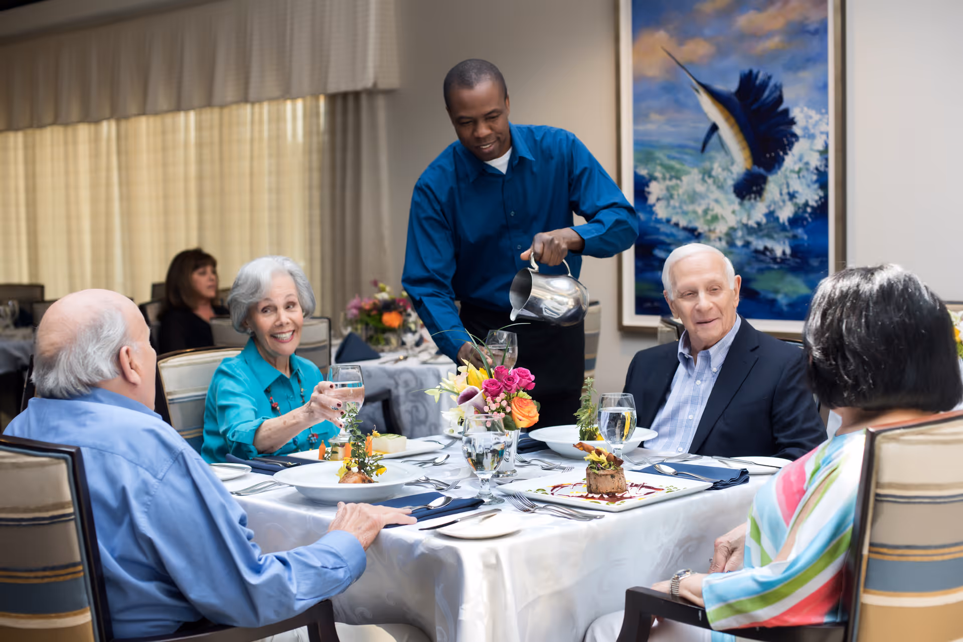 A server pours water for a group of smiling seniors seated around a flower-adorned dining table.