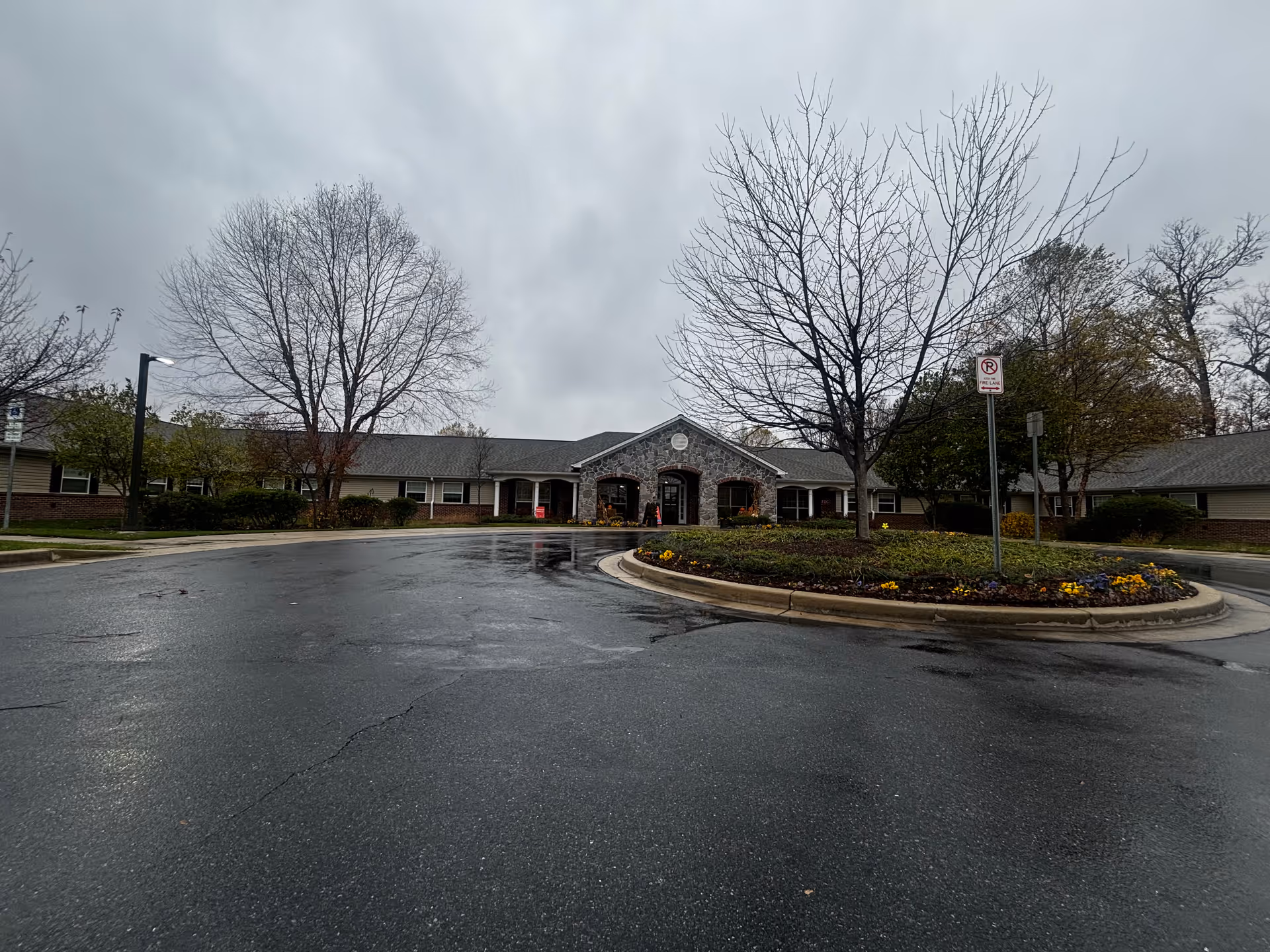 Exterior view of a single-story building with a stone facade entrance, surrounded by leafless trees and a circular driveway with a landscaped island in the center. The sky is overcast and the ground appears wet from recent rain.