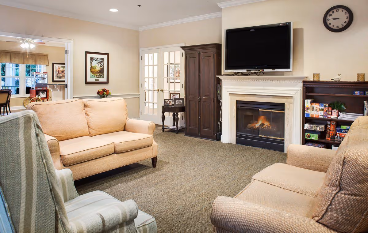 A cozy living room with beige sofas and a striped armchair arranged around a fireplace with a flat-screen TV mounted above it. There is a wooden cabinet and a bookshelf with games and books next to the fireplace. A round clock hangs on the wall above the TV. French doors and a small table with framed photos are visible in the background, along with a glimpse of a dining area.