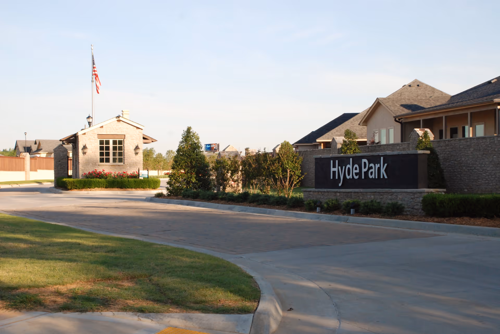 Entrance to Hyde Park at Tulsa Hills senior living facility with a stone guardhouse, an American flag on a flagpole, landscaped greenery, and a large stone sign displaying 'Hyde Park'.