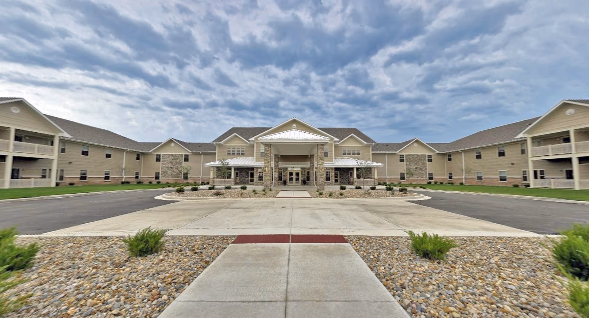 Front entrance of a two-story beige retirement community building with a circular driveway, landscaped walkway, and cloudy sky.
