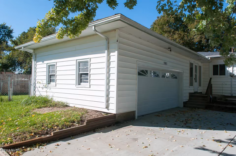 Exterior view of a white single-story building with a garage door numbered 203, a small set of stairs leading to a side entrance, and a green lawn with trees casting shadows.