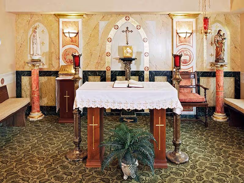 Interior view of a small chapel or prayer room with an altar covered by a white cloth featuring gold crosses. Behind the altar is a decorative wall with religious symbols, a crucifix, and two statues on red marble columns. There are two red candles on ornate candle holders on either side of the altar, a wooden chair with red upholstery, and a green potted plant in front of the altar. The floor is covered with a patterned carpet.