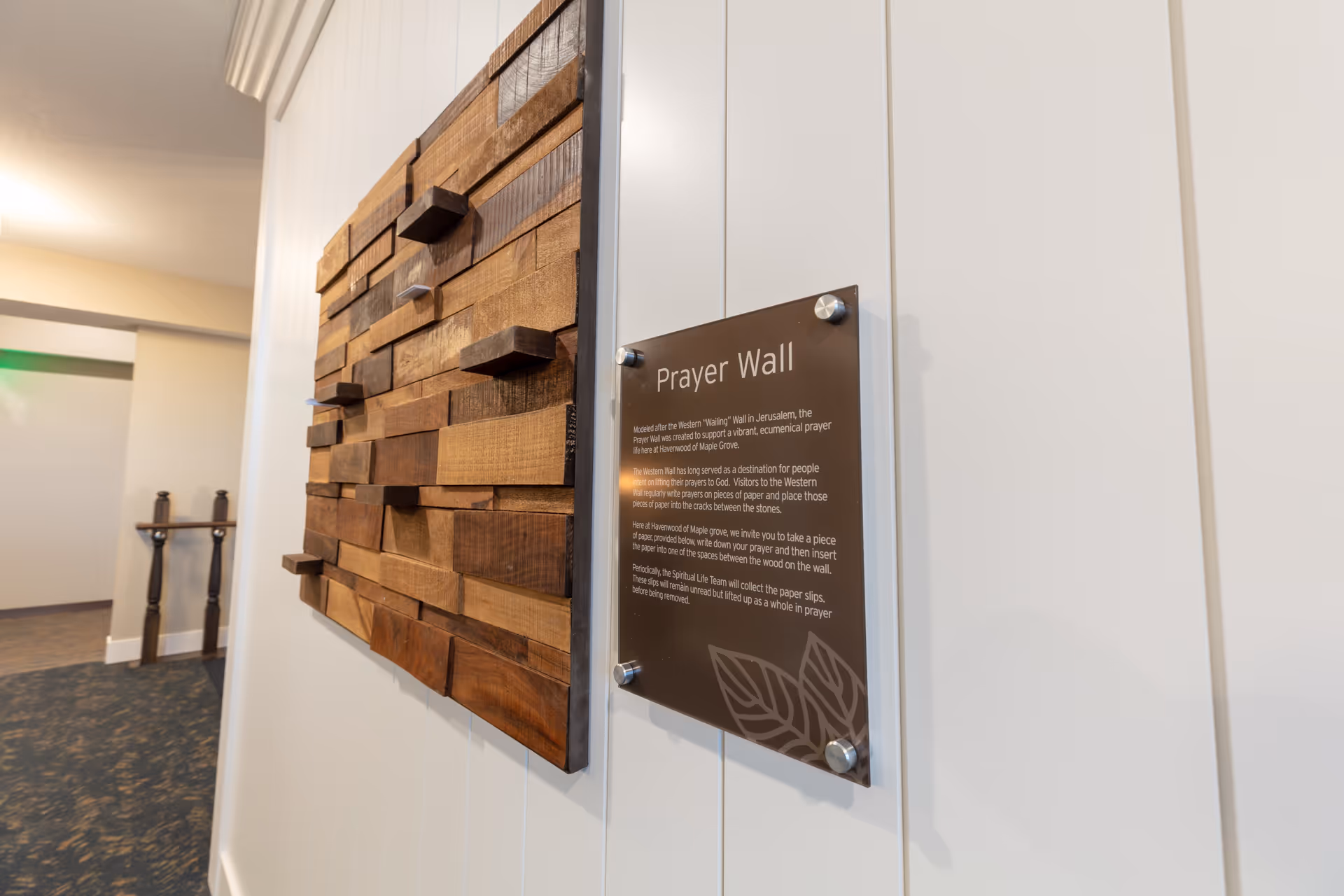 A wooden decorative prayer wall with an informational 'Prayer Wall' plaque mounted on a white paneled hallway wall.
