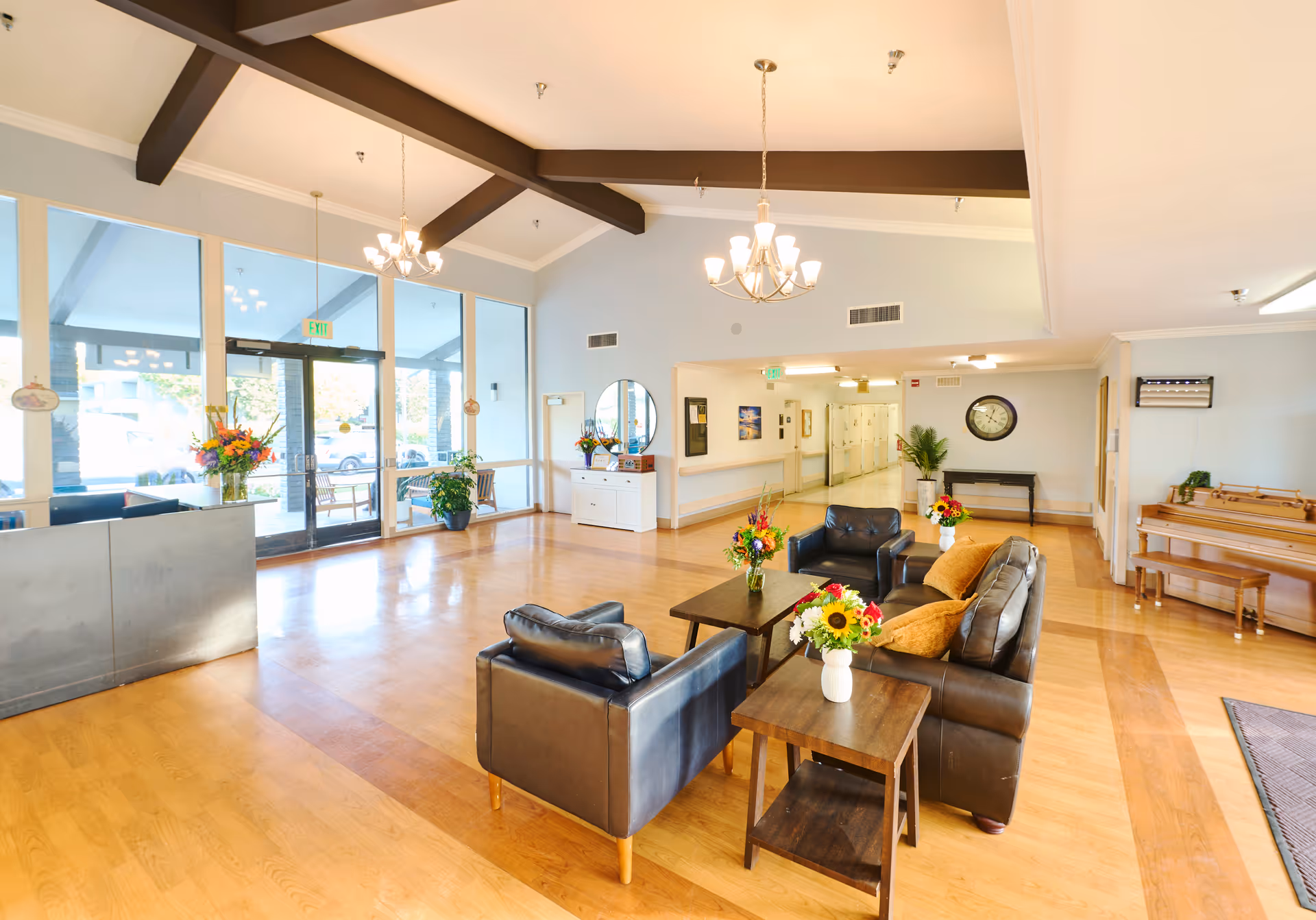 A spacious and bright senior living facility lobby with wooden floors, large windows, and glass doors letting in natural light. The seating area includes black leather chairs and a sofa with yellow cushions around wooden coffee tables decorated with flower vases. There are chandeliers hanging from the ceiling, a piano with a bench on the right side, and a hallway leading further into the building. The entrance area also has plants and a reception desk.