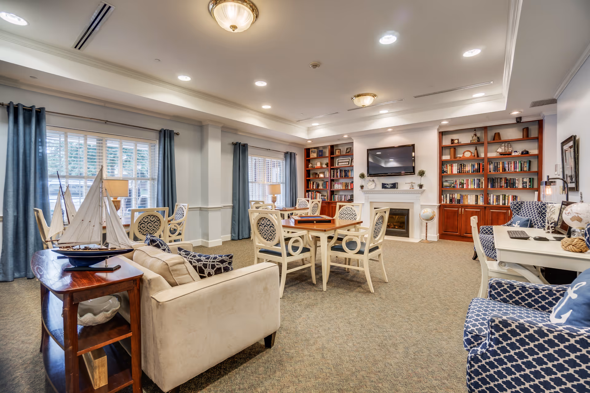 Spacious assisted living lounge with multiple seating areas, tables, built-in bookshelves, a fireplace and wall-mounted TV.