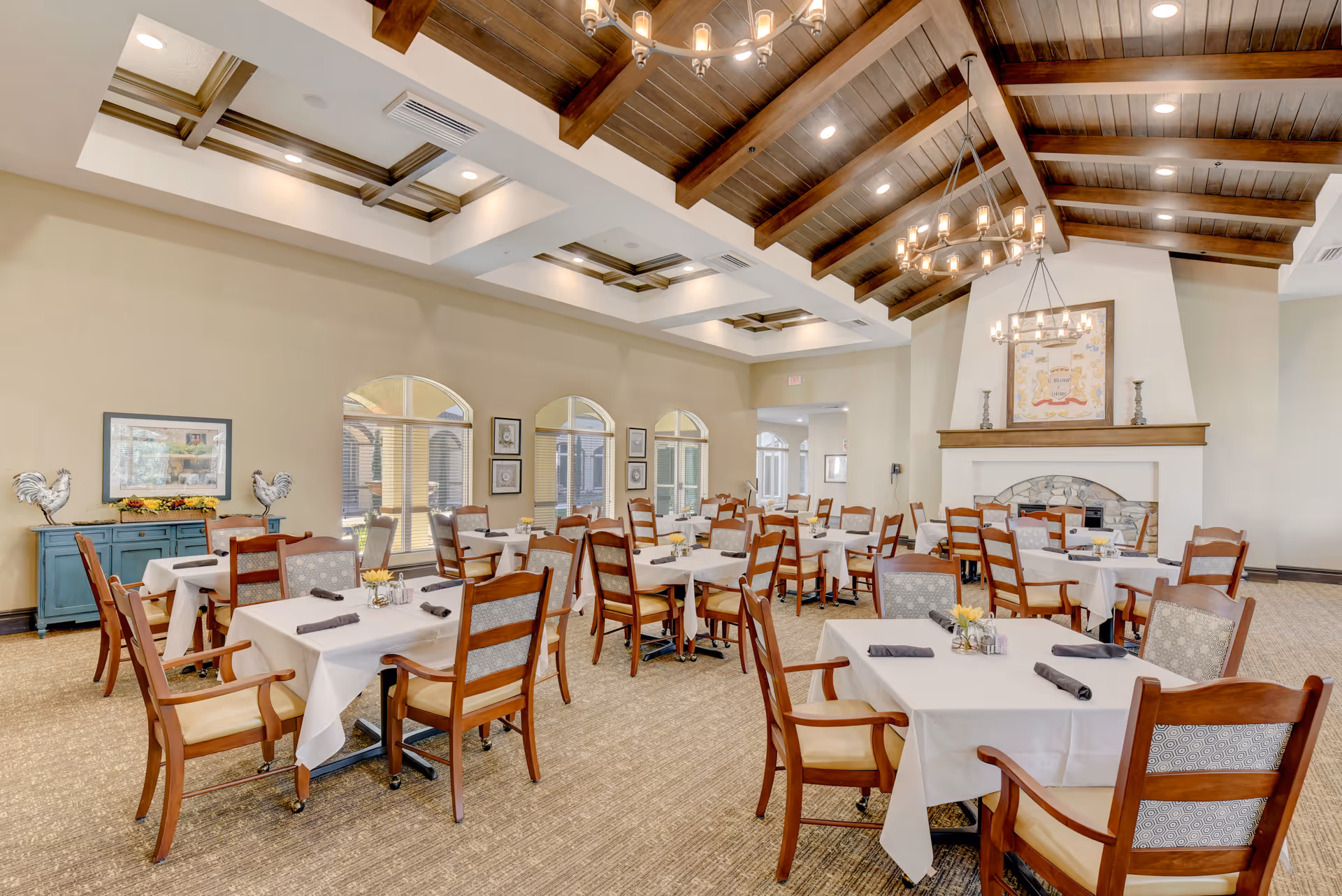A spacious dining room with multiple tables covered in white tablecloths, each set with napkins and small flower arrangements. The room features wooden chairs with cushioned seats, a high ceiling with exposed wooden beams, and two chandeliers. There is a fireplace with a framed artwork above it and several windows along one wall letting in natural light.
