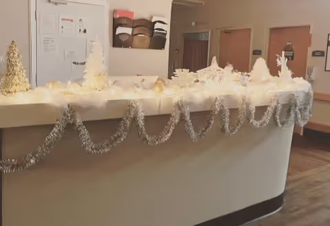 Reception desk decorated with white and silver holiday decorations including garlands, small trees, and lights in a healthcare facility hallway with doors and bulletin boards in the background.