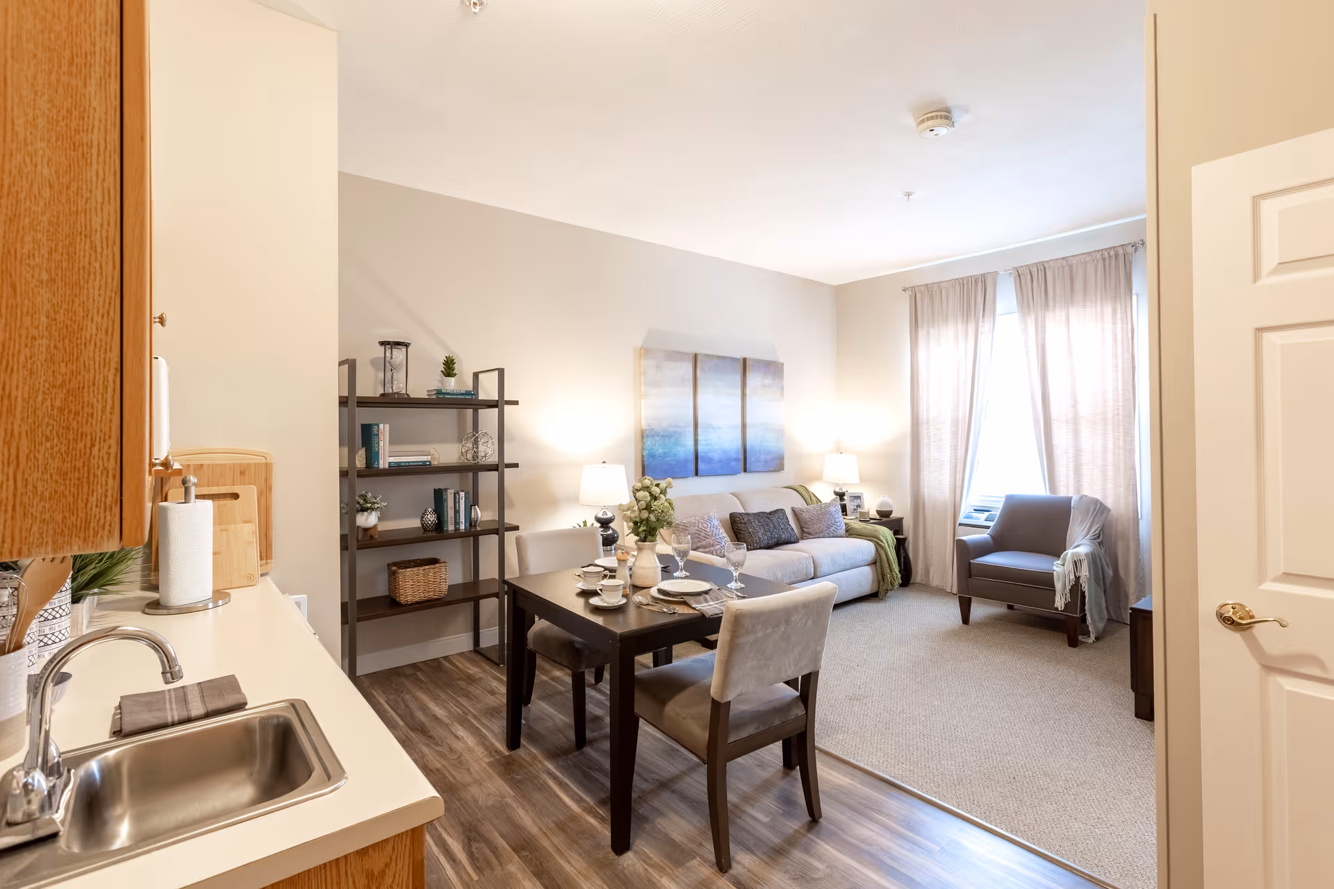 A bright and cozy living space in an apartment at Independence Village of Fishers South featuring a small kitchen area with a sink and wooden cabinets on the left, a dining table set for four in the center, and a living room area with a sofa, armchair, side tables with lamps, and a window with curtains letting in natural light. A bookshelf with decorative items is against the wall.