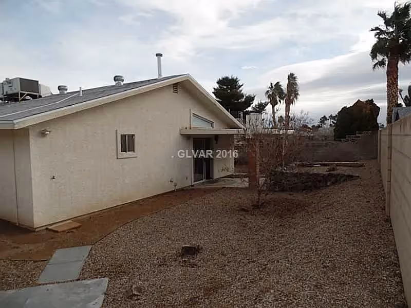 Backyard of a single-story beige house with gravel ground, a sliding patio door, and palm trees beyond a block wall.