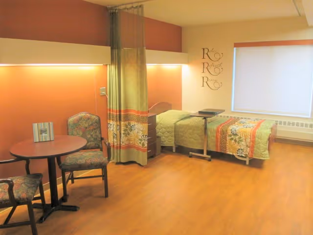 A nursing facility patient room with a single bed, privacy curtain, bedside table, round dining table with two chairs, and wood-look flooring.