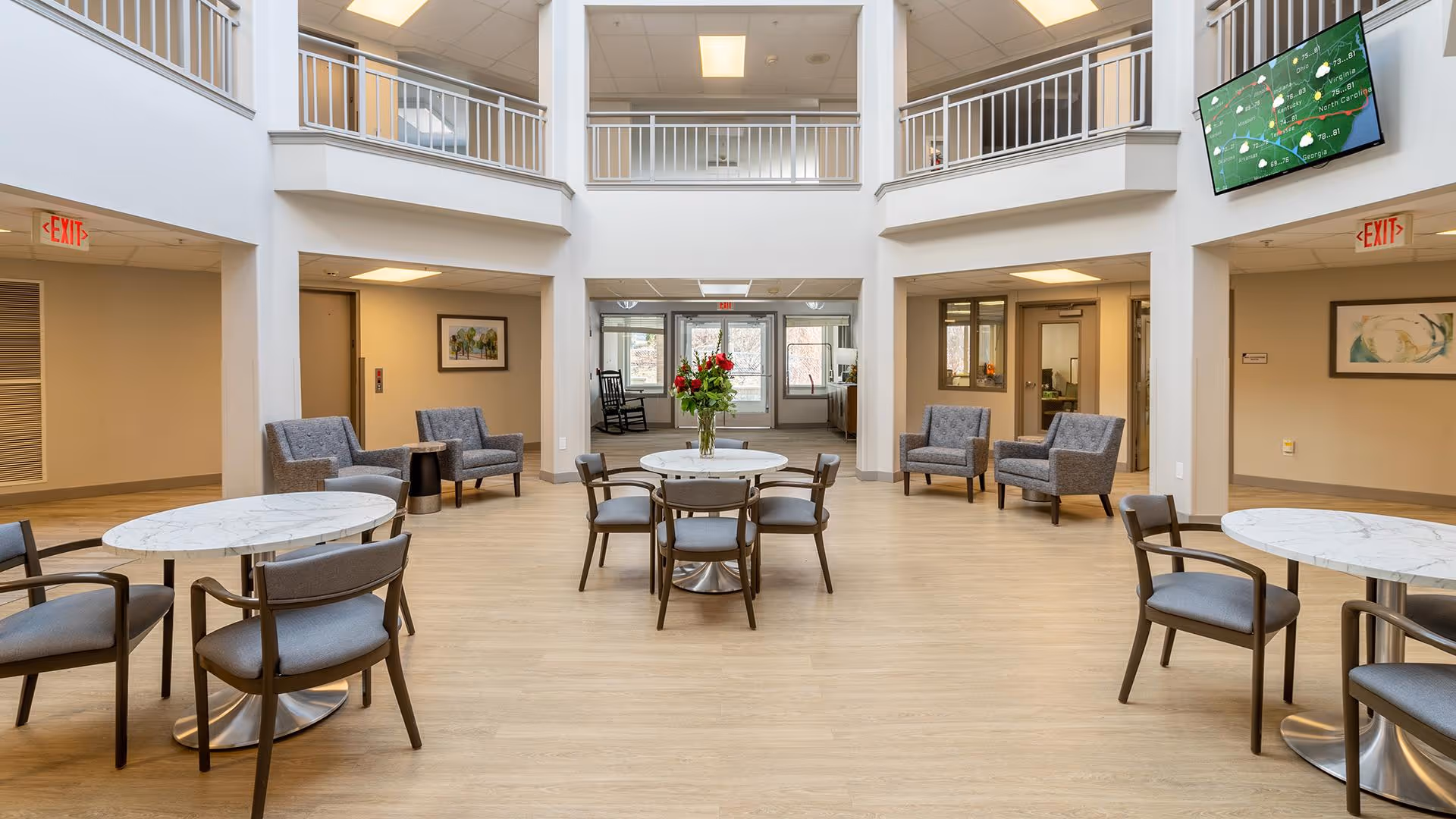 A spacious, well-lit common area in a senior living facility with round tables and chairs arranged for seating. There are several armchairs along the walls, a vase with flowers on the central table, and a large TV mounted on the upper right wall. The area has a high ceiling with a balcony railing above and exit signs visible on both sides.