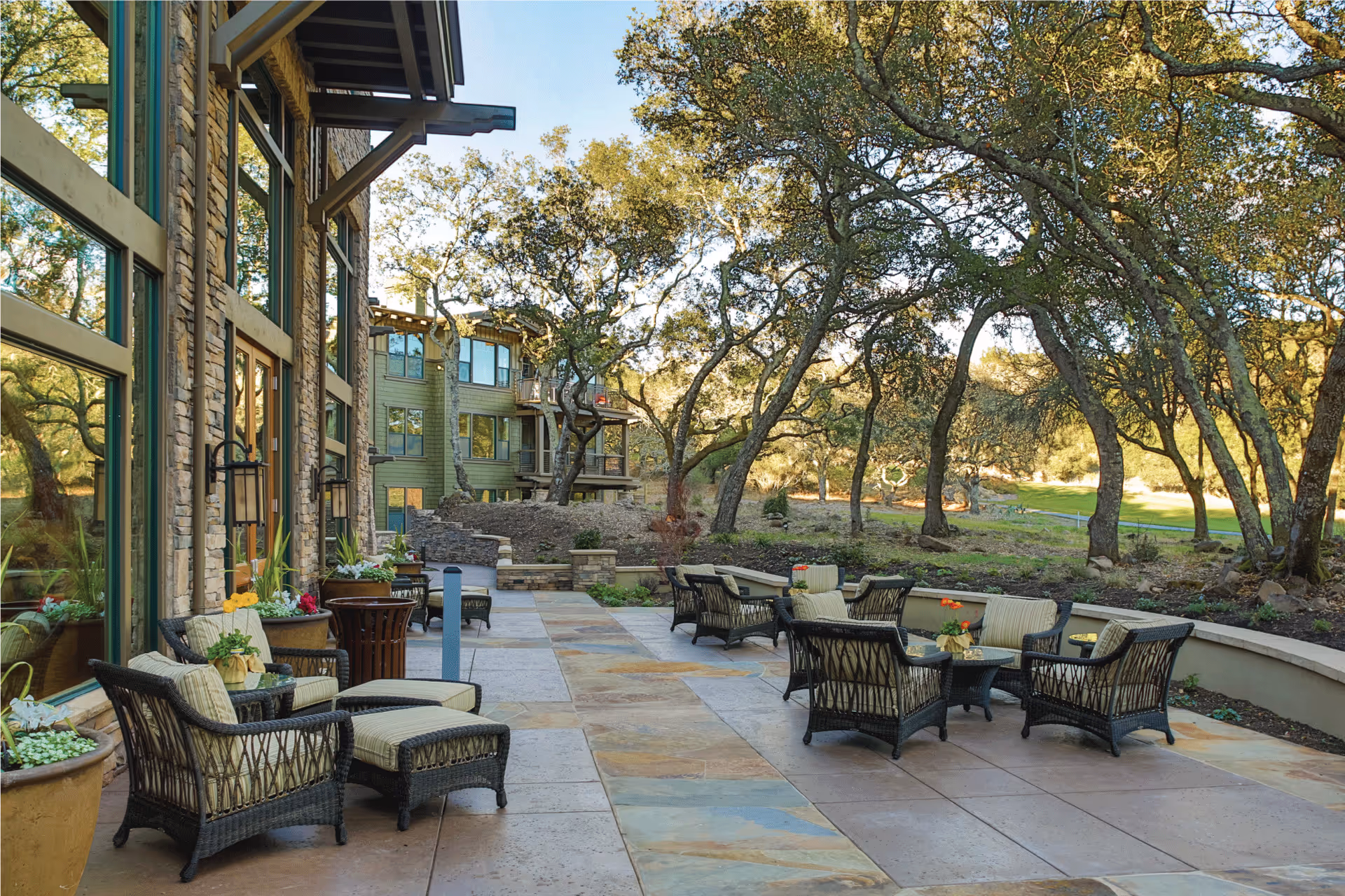 Outdoor patio area at Fountaingrove Lodge with wicker chairs and tables arranged on a stone-tiled floor. The patio is adjacent to a building with large windows and stone walls, surrounded by trees and greenery under a clear sky.