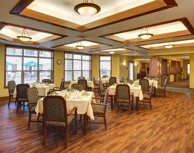 A spacious and well-lit dining room in a senior living facility with multiple tables covered in white tablecloths, each set with napkins, glasses, and silverware. The room features large windows allowing natural light, wooden flooring, and ceiling lights with decorative wooden beams.