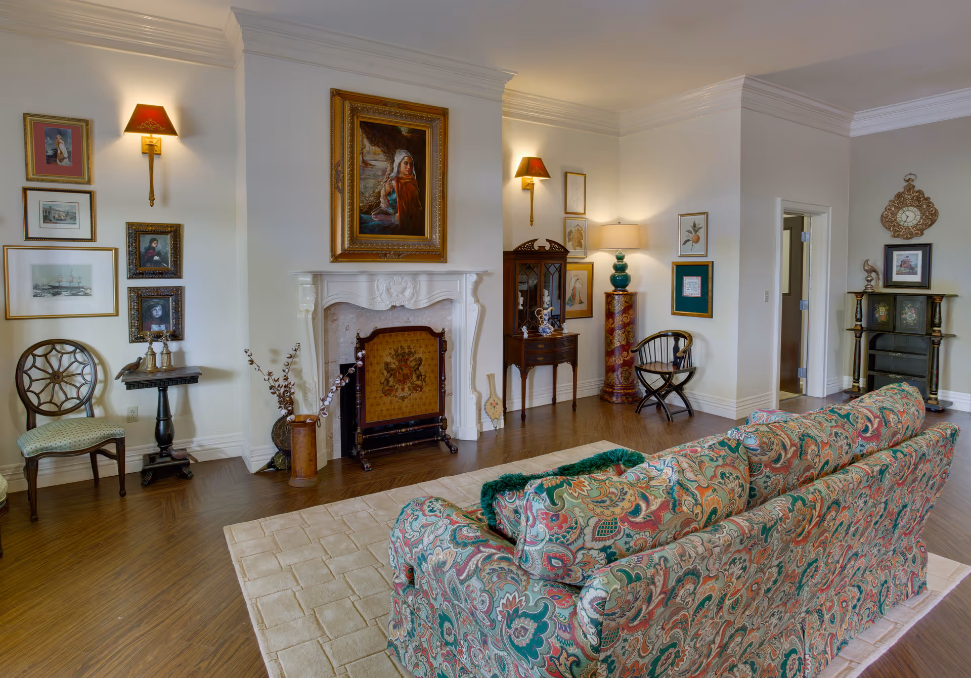 A cozy living room with a patterned sofa facing a white ornate fireplace. The walls are decorated with framed paintings and pictures. There are two chairs, a small table, and a decorative cabinet. The room has wooden flooring and soft lighting from wall sconces and a table lamp.