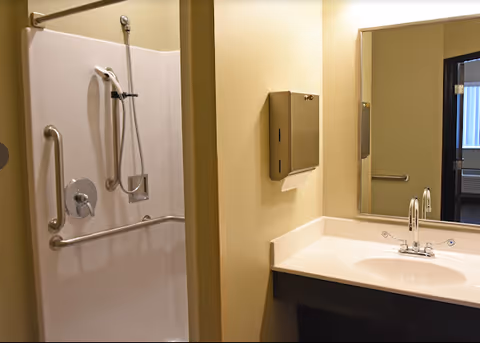 Accessible bathroom with a walk-in shower with grab bars, handheld showerhead, and a sink with mirror and paper towel dispenser.