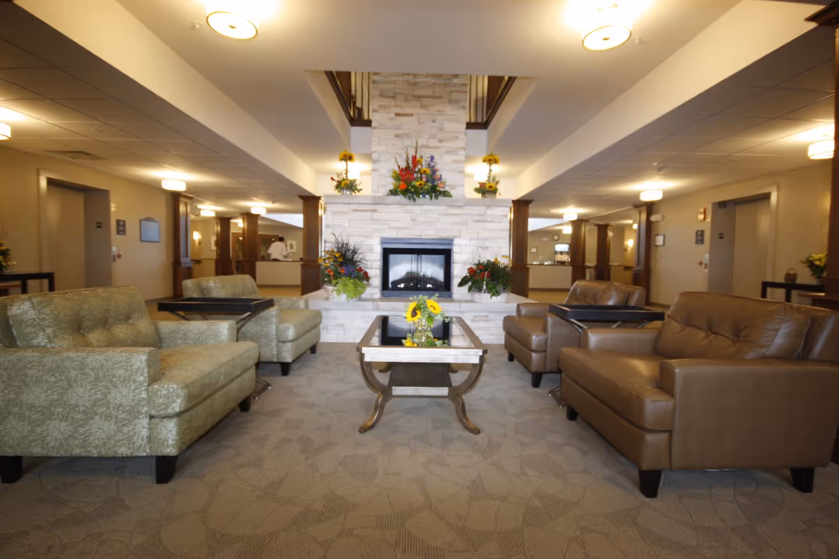 A spacious and well-lit common area with a central stone fireplace adorned with colorful flower arrangements. The room features comfortable seating including two green patterned armchairs and two brown leather armchairs arranged around a glass coffee table with a small vase of sunflowers. The background shows a reception desk and hallways with warm lighting and wooden columns.