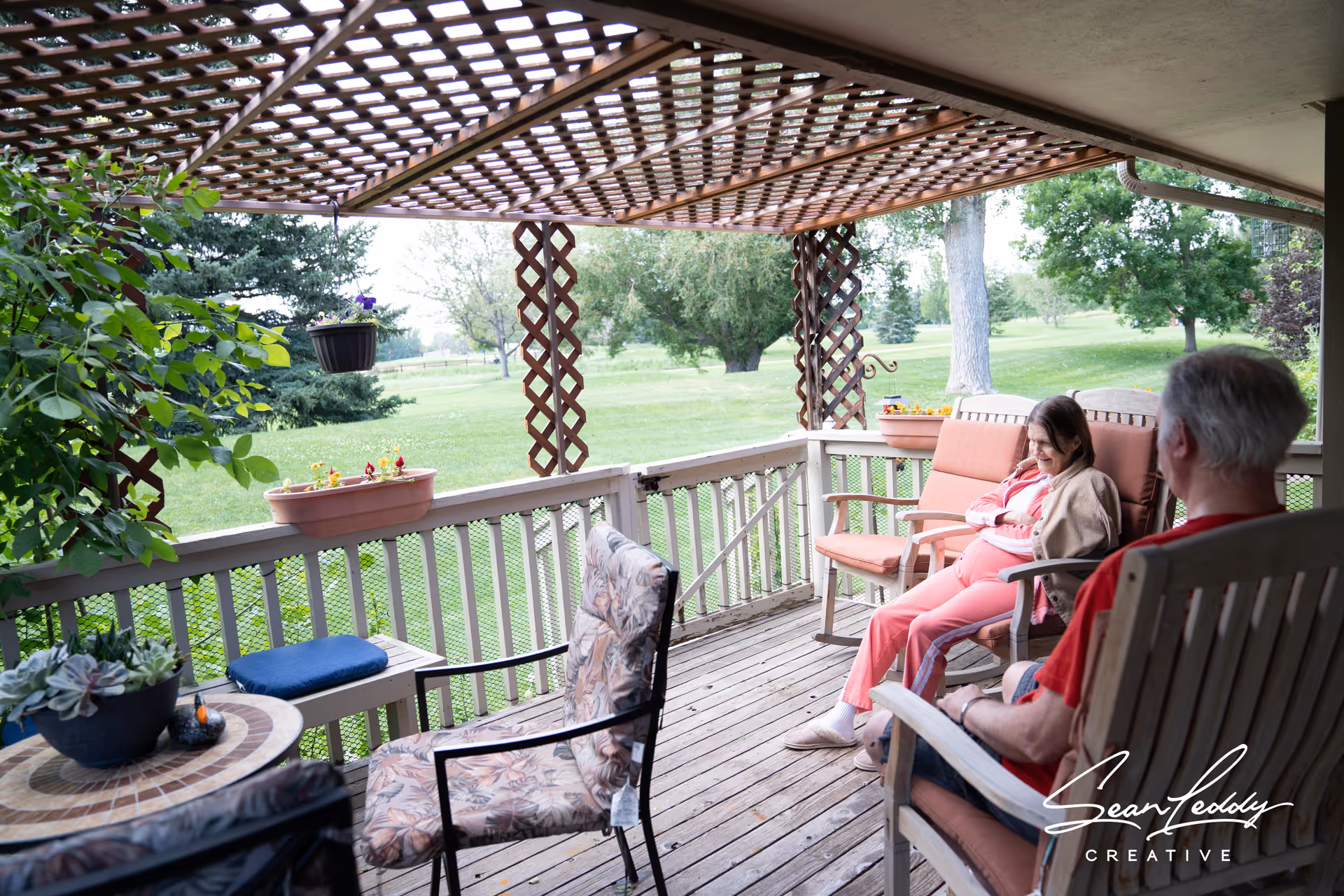 Two seniors sitting on a covered wooden porch overlooking a grassy yard with trees.