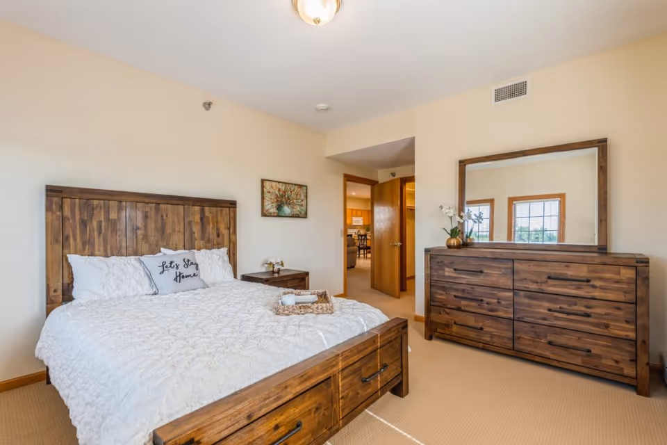 Bright furnished bedroom with a wooden bed and matching dresser, white bedding and a decorative pillow, and an open doorway to another room.